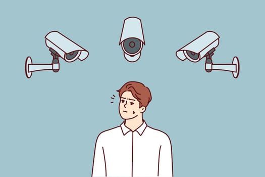 Embarrassed man stands among cameras recording every step of city dwellers to prevent commission of crimes. Dissatisfied man looking at CCTV cameras wondering if Big Brother is spying