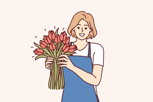 Woman flower seller stands with luxurious bouquet and smiling looks at camera enjoying work of florist in flower shop. Positive girl in apron holds fragrant bouquet with scarlet buds