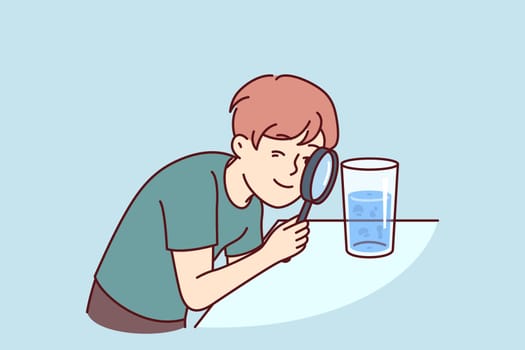 Small boy examines water in glass through magnifying glass, studying chemical composition liquid or looking for microbes. Teenage child curiously studies water, wanting to work in chemical laboratory