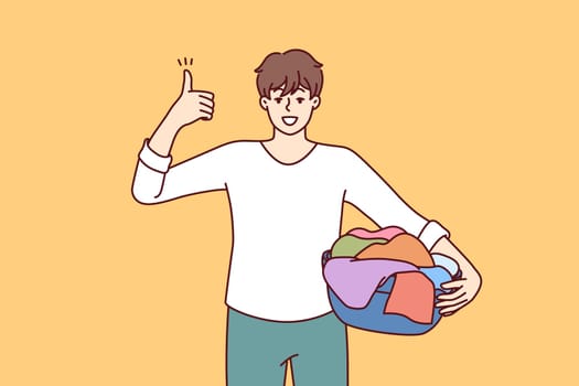 Man householder with laundry in basin shows thumbs up recommending quality laundry detergent. Guy getting ready to load textiles into washing machine setting positive example for peers