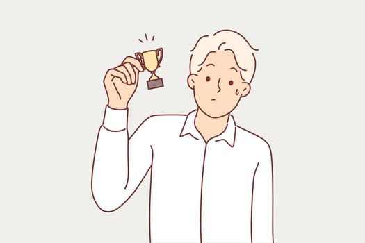 Man with small cup for minor achievement feels displaced and awkward after receiving consolation prize. Loser guy demonstrates miniature golden trophy symbolizing fraud in prize draw