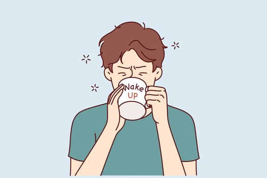 Man drinks coffee from mug with inscription wake up, wanting to cheer up and gain strength before difficult working day. Tired young guy trying to wake up with caffeine and taurine drinks